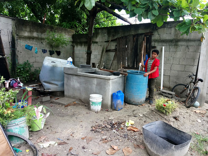 Recipientes de almacenamiento de agua (de izquierda a derecha): cisterna (con la botella de cloro en la parte superior), pila (el depósito de cemento), cubeta, barril plástico. La ropa se lava en tablas de lavar como se ve al lado izquierdo de la pila. El muchacho es un voluntario de Monte Verde, comprobando para larvas de mosquitos en el barril.